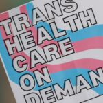 The pink, blue and white striped symbol of the transgender pride flag reads: "Trans health care on demand!"